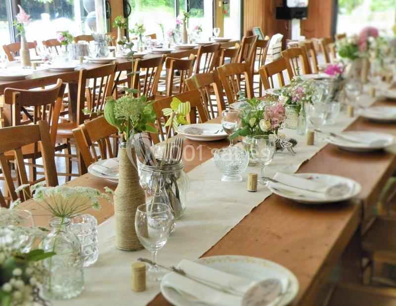 Tables en bois décorées de fleurs, vaisselle et verres, préparées pour un repas dans une salle lumineuse.