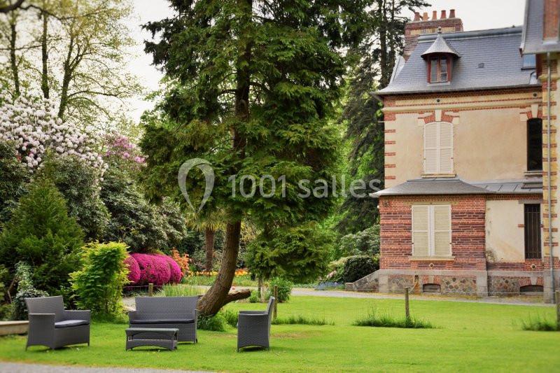 Mobilier de jardin en rotin noir disposé sur une pelouse, près d'un arbre et d'une maison en briques avec volets fermés.