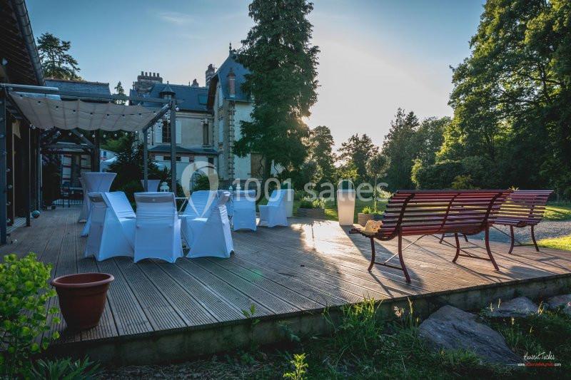 Terrasse en bois avec chaises blanches, bancs et vue sur un jardin arboré au coucher du soleil.