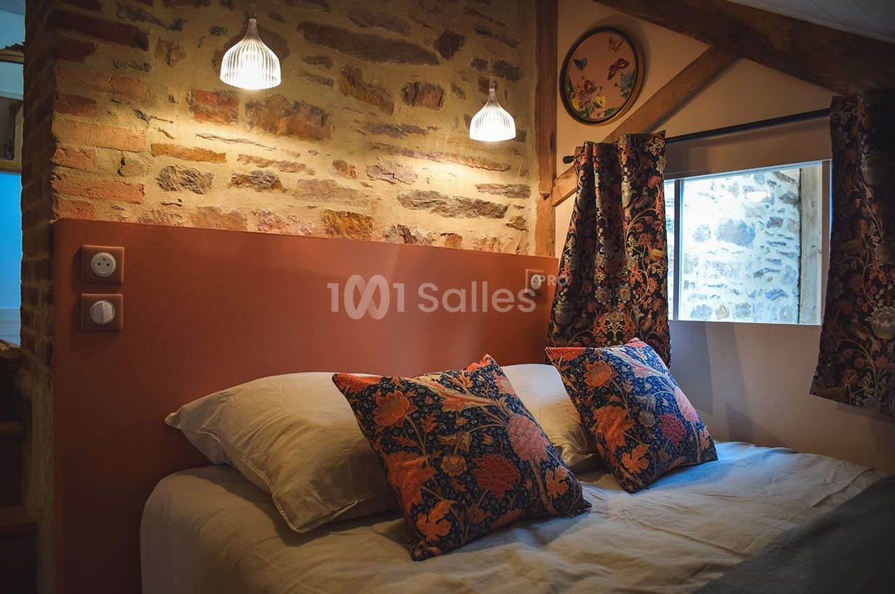 Lit double avec coussins à motifs floraux, éclairé par des lampes suspendues, dans une chambre aux murs en pierre apparente.