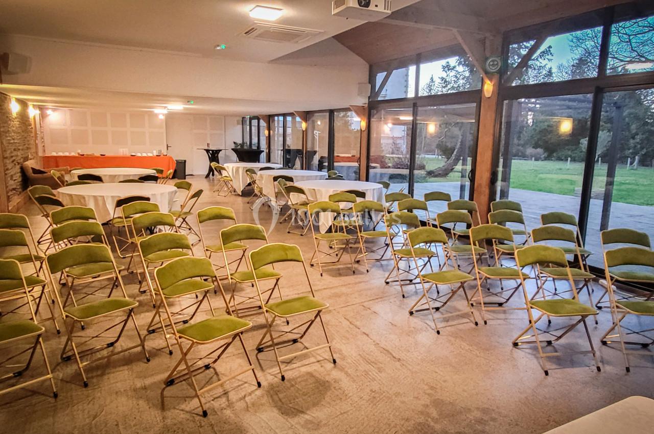 Salle lumineuse avec chaises pliantes vertes disposées face à des tables, grandes baies vitrées donnant sur un espace…