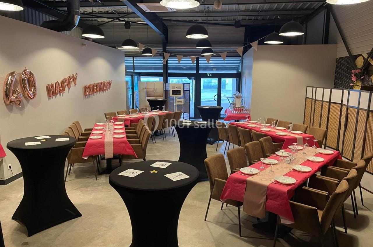 Salle décorée pour une fête d'anniversaire avec tables dressées, nappes roses et ballons ’Happy Birthday’.