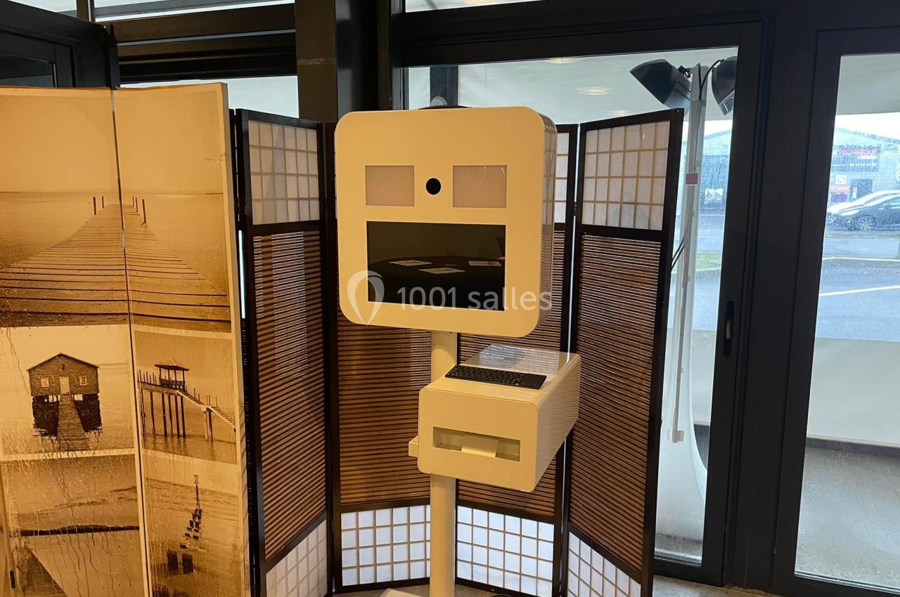 Borne photo moderne sur pied, placée devant un paravent en bois et papier, dans un espace intérieur lumineux.