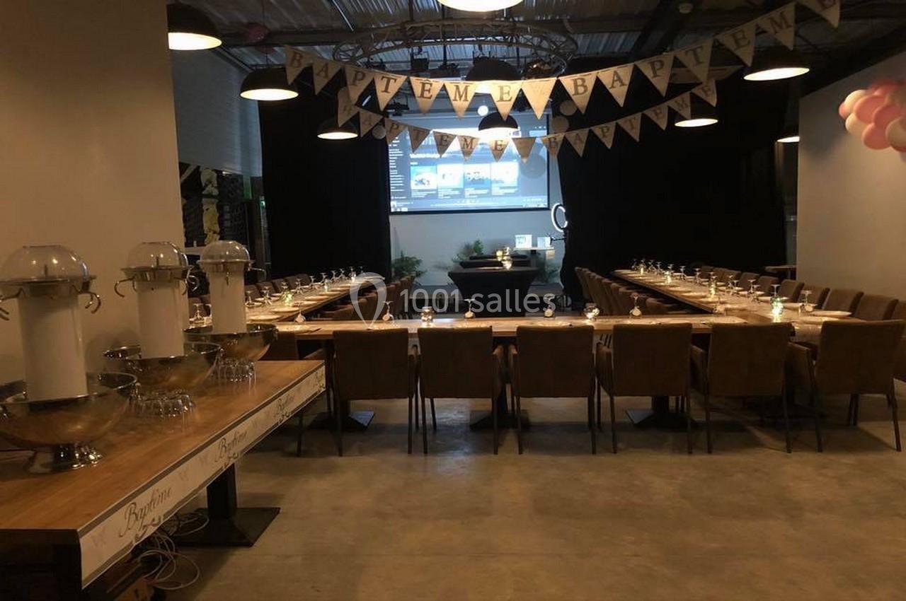 Salle décorée pour un baptême avec tables alignées, chaises en cuir, guirlandes et distributeurs de boissons.