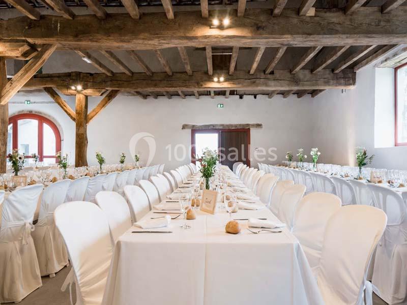 Salle de réception avec longues tables dressées, nappes blanches, chaises houssées et poutres apparentes au plafond.