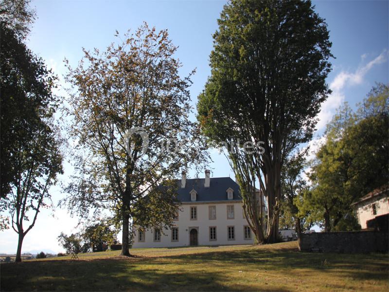 Grande maison blanche entourée d'arbres dans un paysage ensoleillé avec pelouse et ciel dégagé.