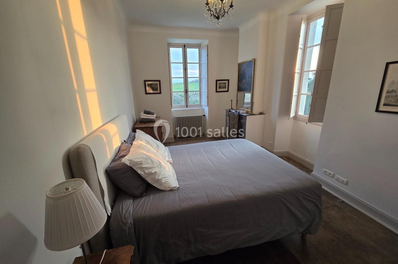 Chambre lumineuse avec un lit double, draps gris, lampes de chevet et fenêtres laissant entrer la lumière du jour.