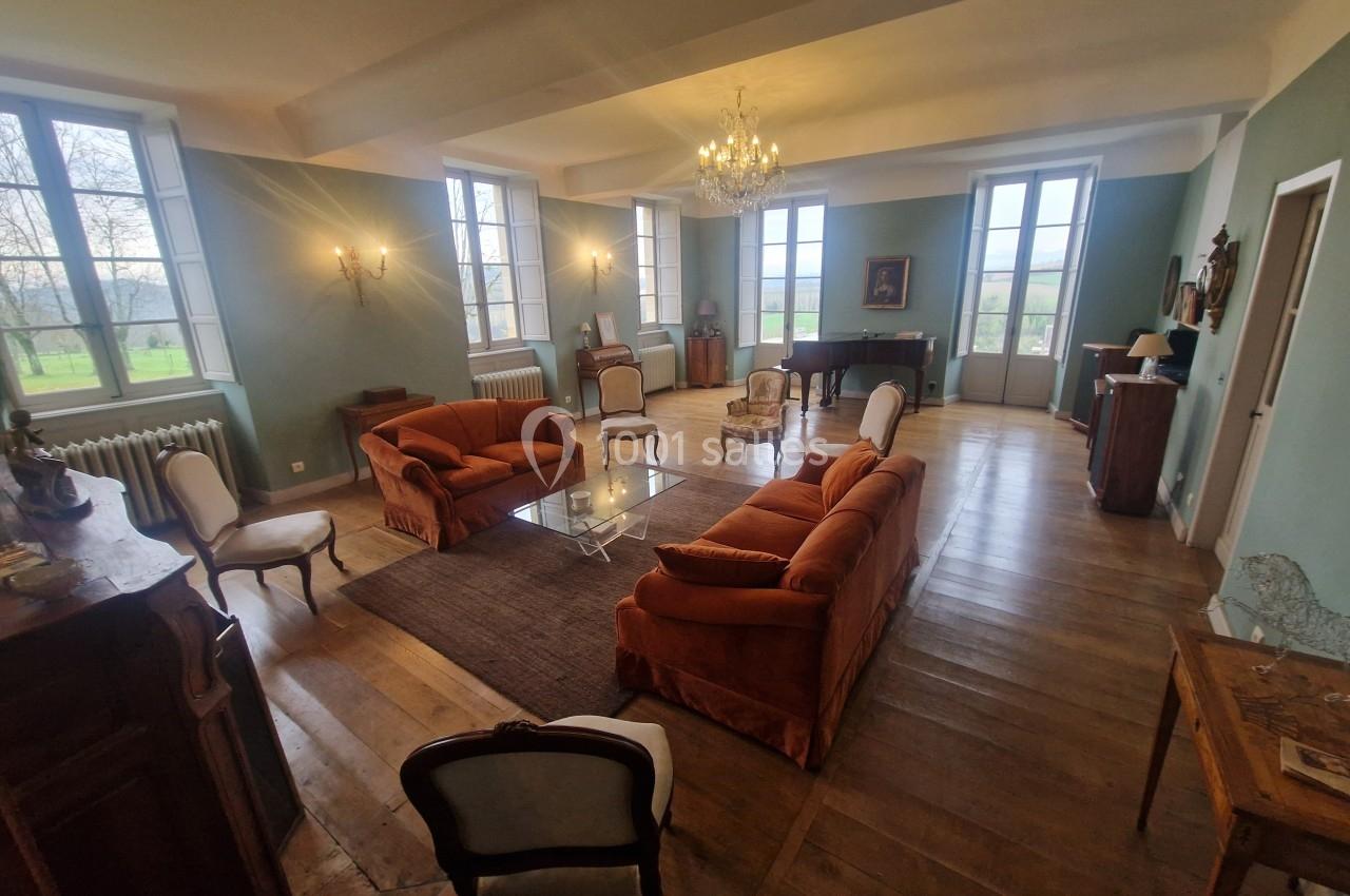 Salon spacieux avec parquet en bois, canapés orange, fauteuils, piano à queue et grandes fenêtres donnant sur un jardin.