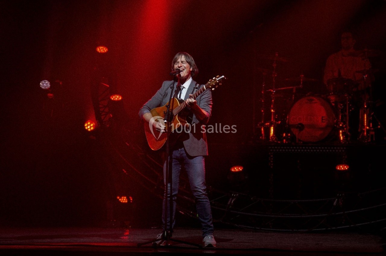 Location salle Dammarie-les-Lys (Seine-et-Marne) - Espace Pierre Bachelet #15 Un musicien joue de la guitare sur une scène éclairée de lumières rouges, avec un batteur en arrière-plan.
