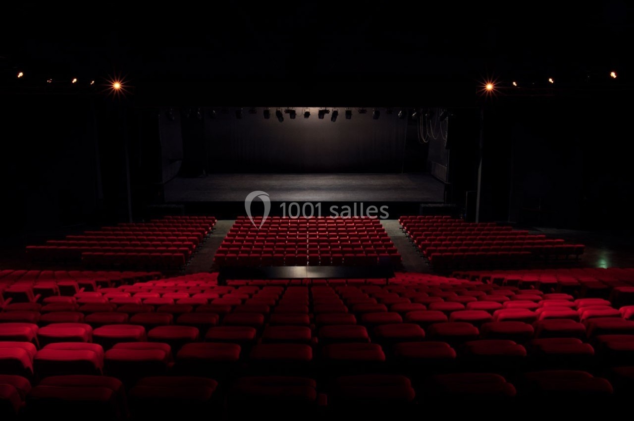 Salle de théâtre vide avec des rangées de sièges rouges et une scène éclairée en arrière-plan.