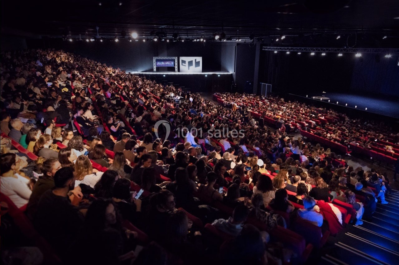 Salle de spectacle bondée avec des spectateurs assis sur des rangées de sièges rouges, face à une scène éclairée.