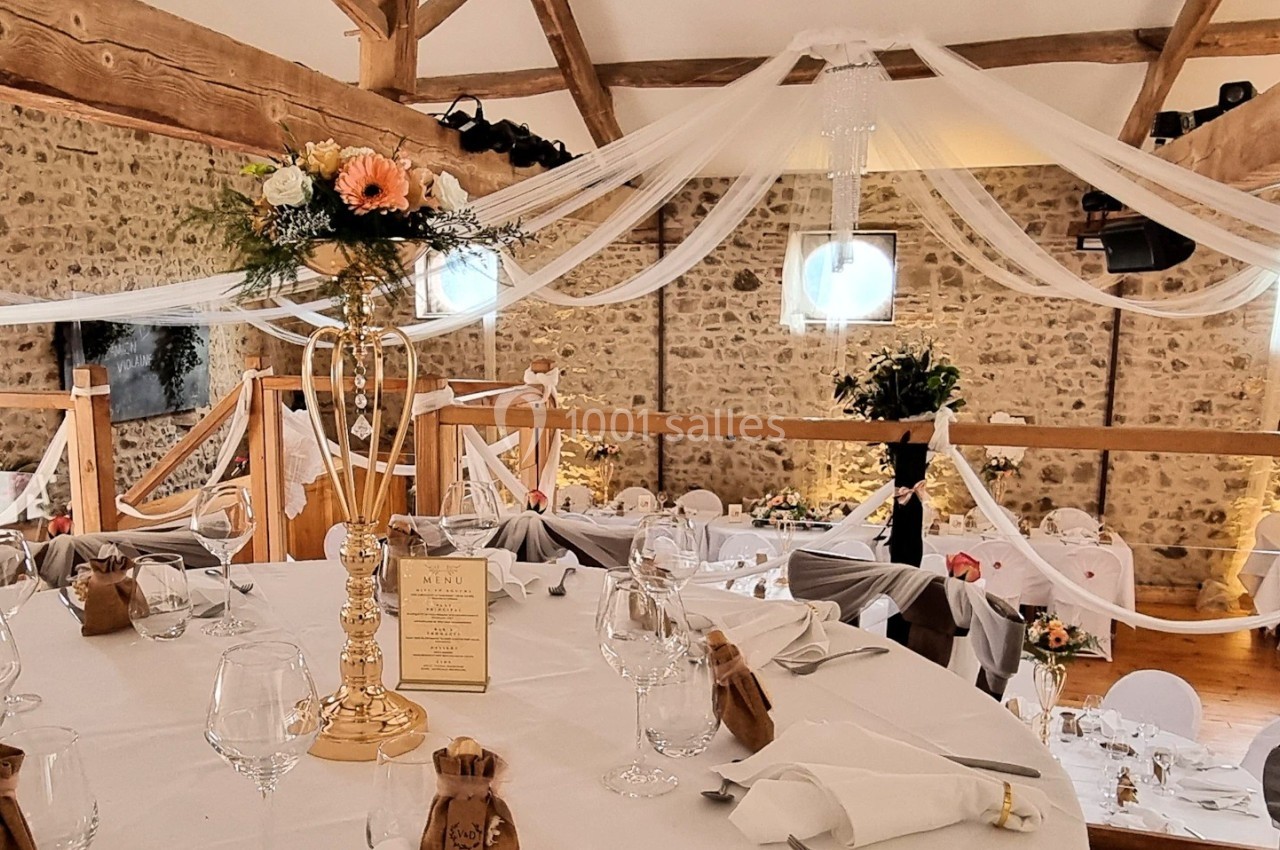 Salle de réception décorée avec des tables dressées, des fleurs et des voilages suspendus sous une charpente en bois.