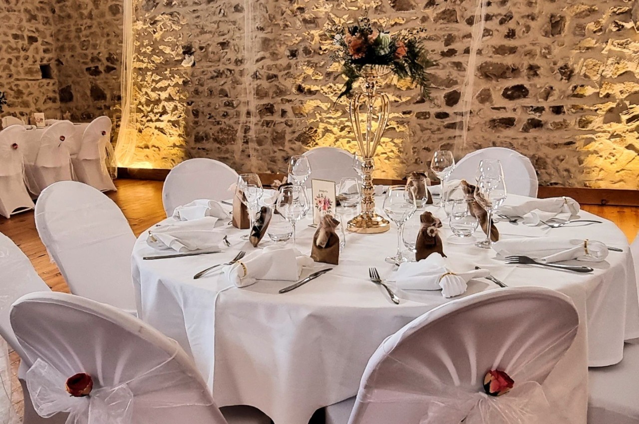 Table ronde décorée pour un mariage, avec nappes blanches, chaises houssées, centre de table floral et murs en pierre…