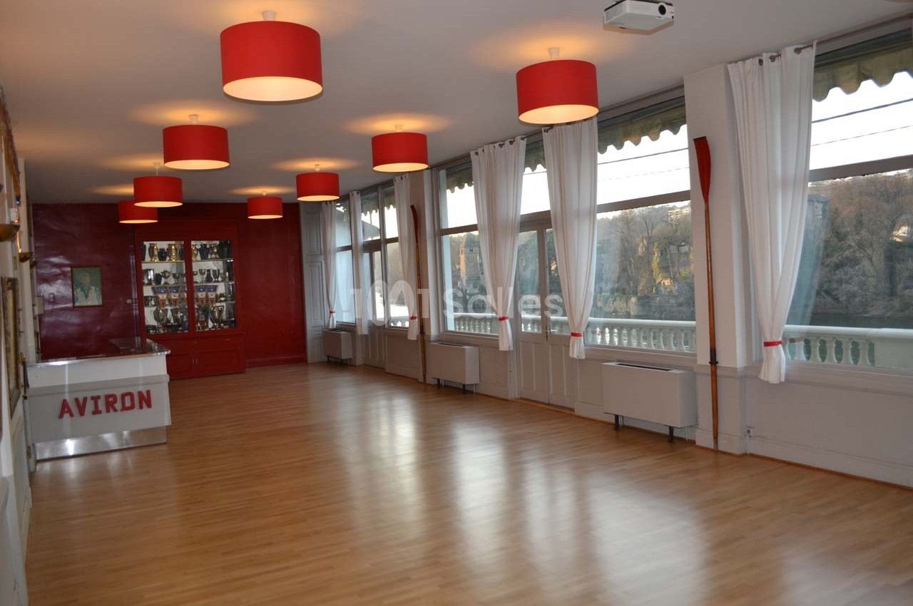 Salle lumineuse avec parquet, grandes fenêtres donnant sur l'extérieur, rideaux blancs et luminaires rouges.