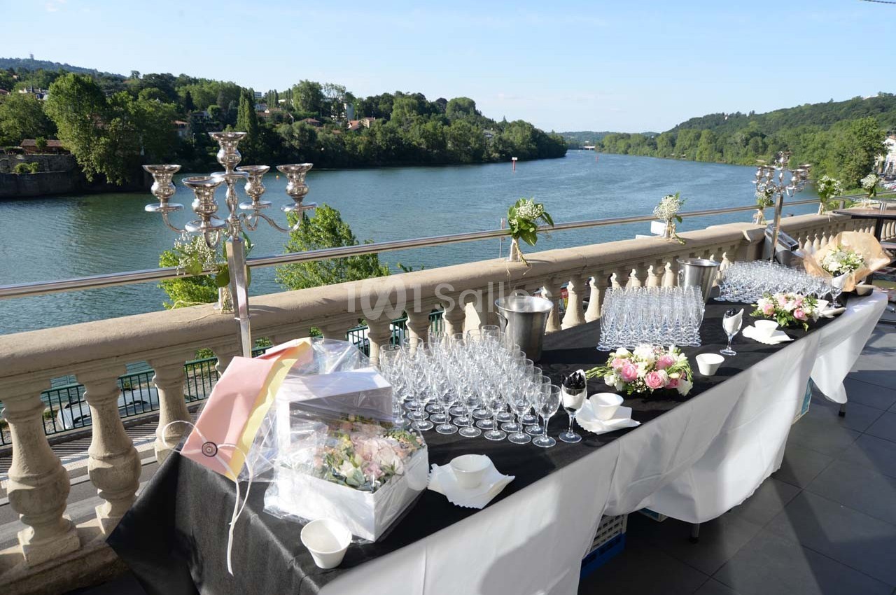 Table dressée avec des verres et des décorations florales sur une terrasse surplombant une rivière et un paysage verdoyant.