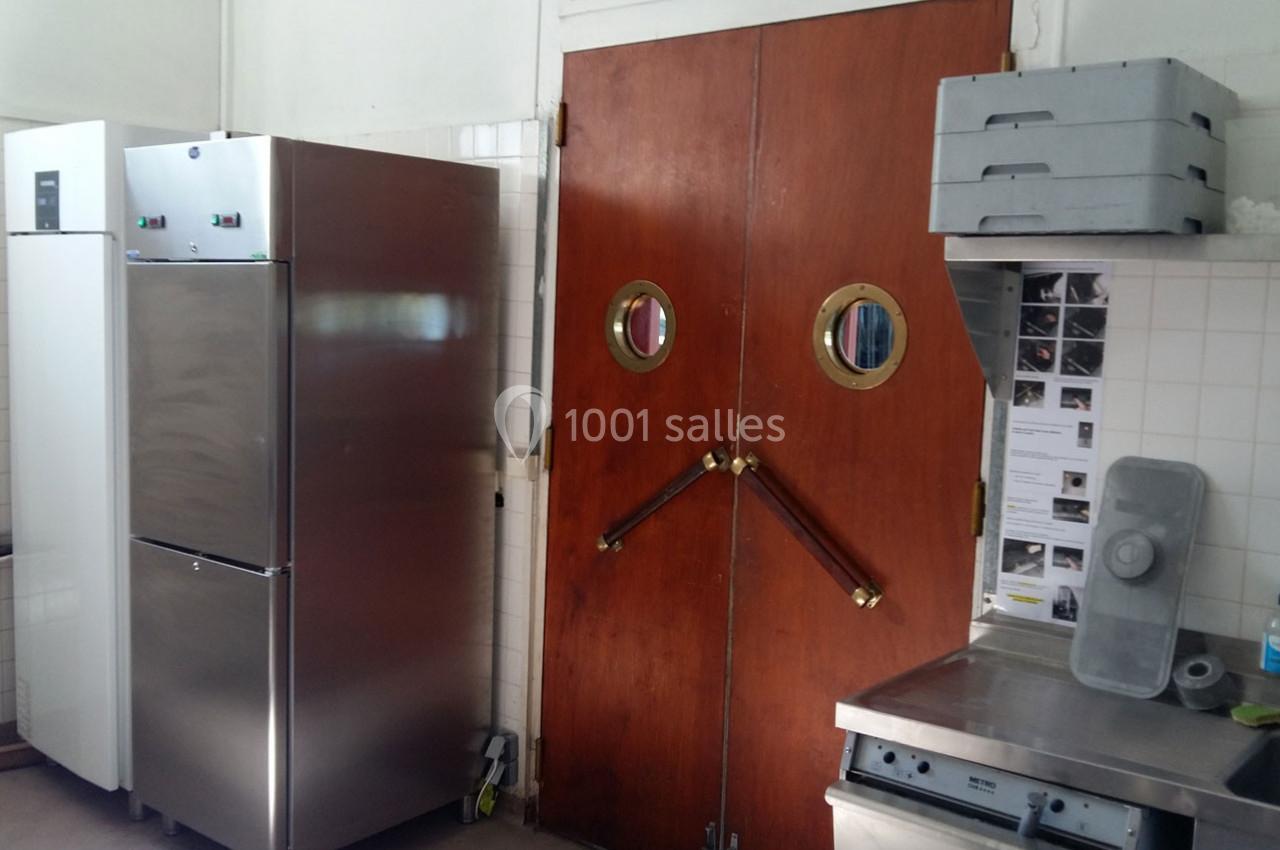 Cuisine professionnelle avec réfrigérateur en acier inoxydable, portes battantes en bois et plans de travail en métal.