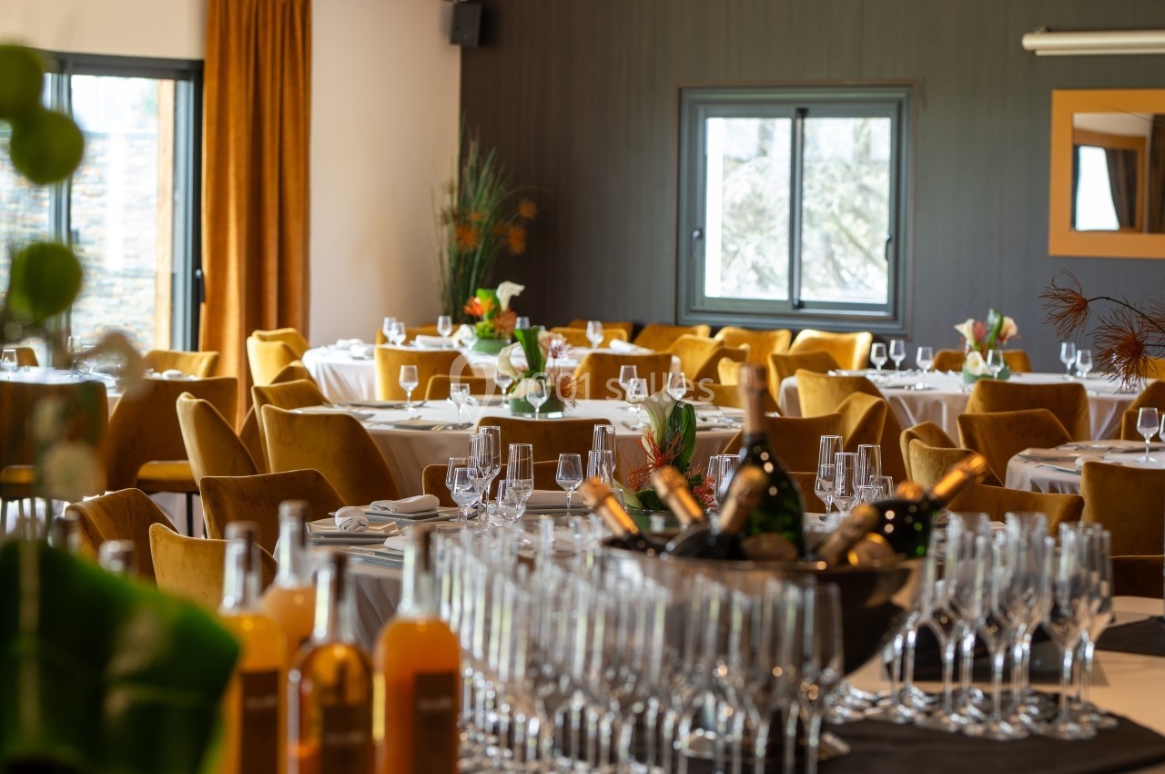 Salle de réception élégante avec tables dressées, chaises jaunes, décor floral et bar avec bouteilles et verres.