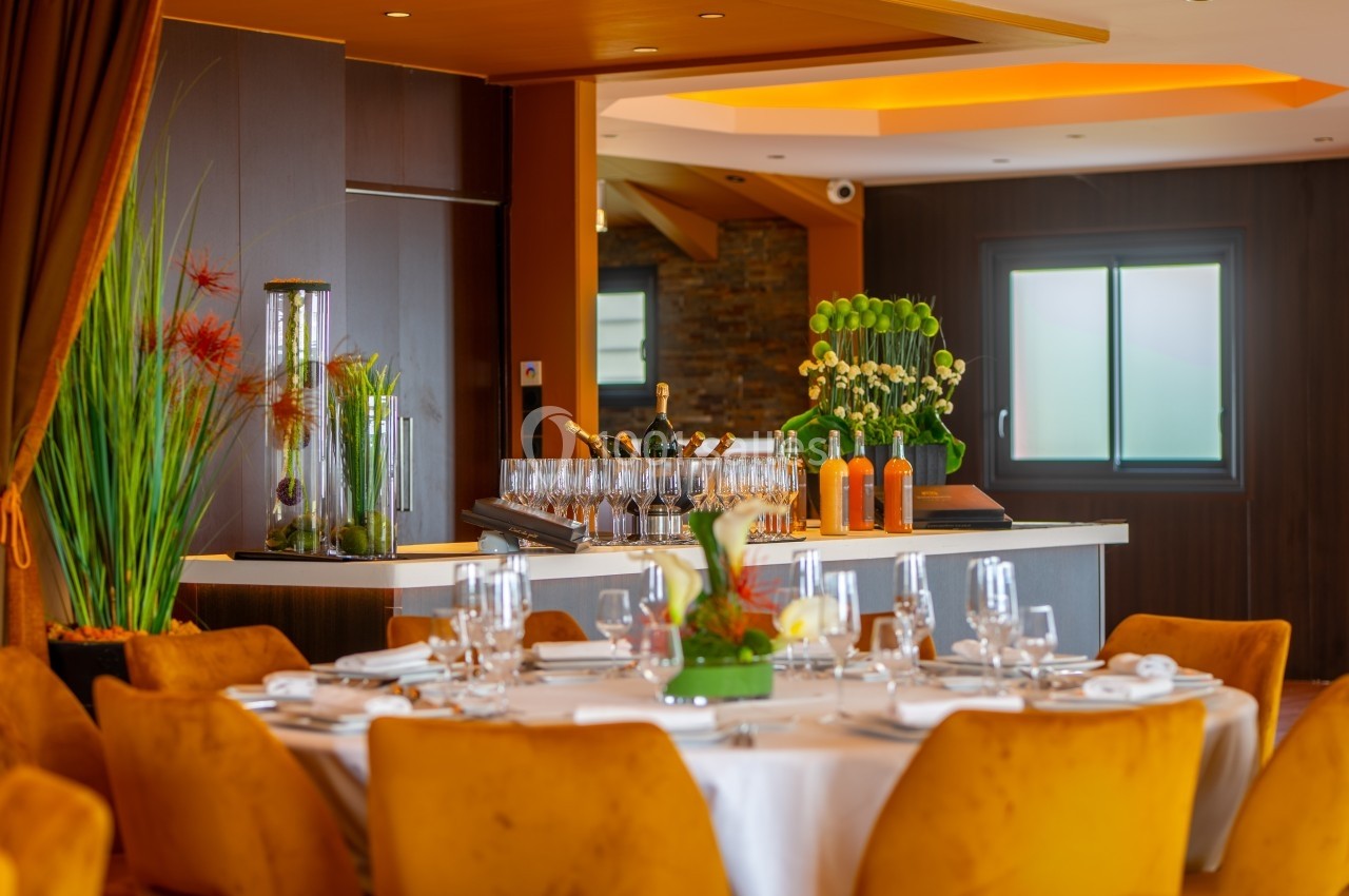 Salle de restaurant élégante avec table dressée, chaises jaunes et bar en arrière-plan décoré de plantes et bouteilles.