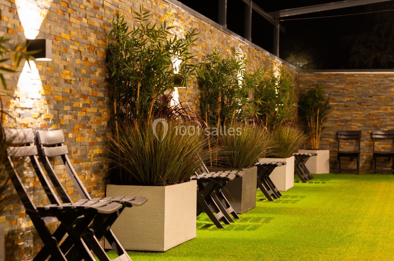 Chaises pliantes noires alignées près de jardinières sur une pelouse artificielle, éclairées par des appliques murales.