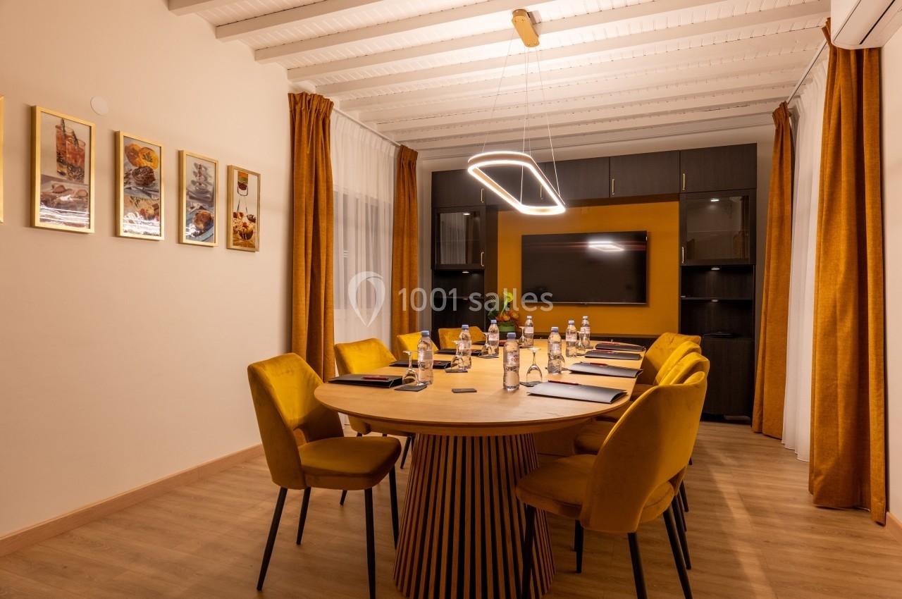 Salle de réunion avec table ronde, chaises jaunes, écran mural, étagères et éclairage suspendu moderne.
