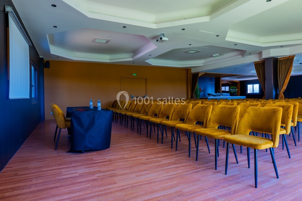 Salle de conférence vide avec rangées de chaises jaunes, table noire et écran de projection au mur.