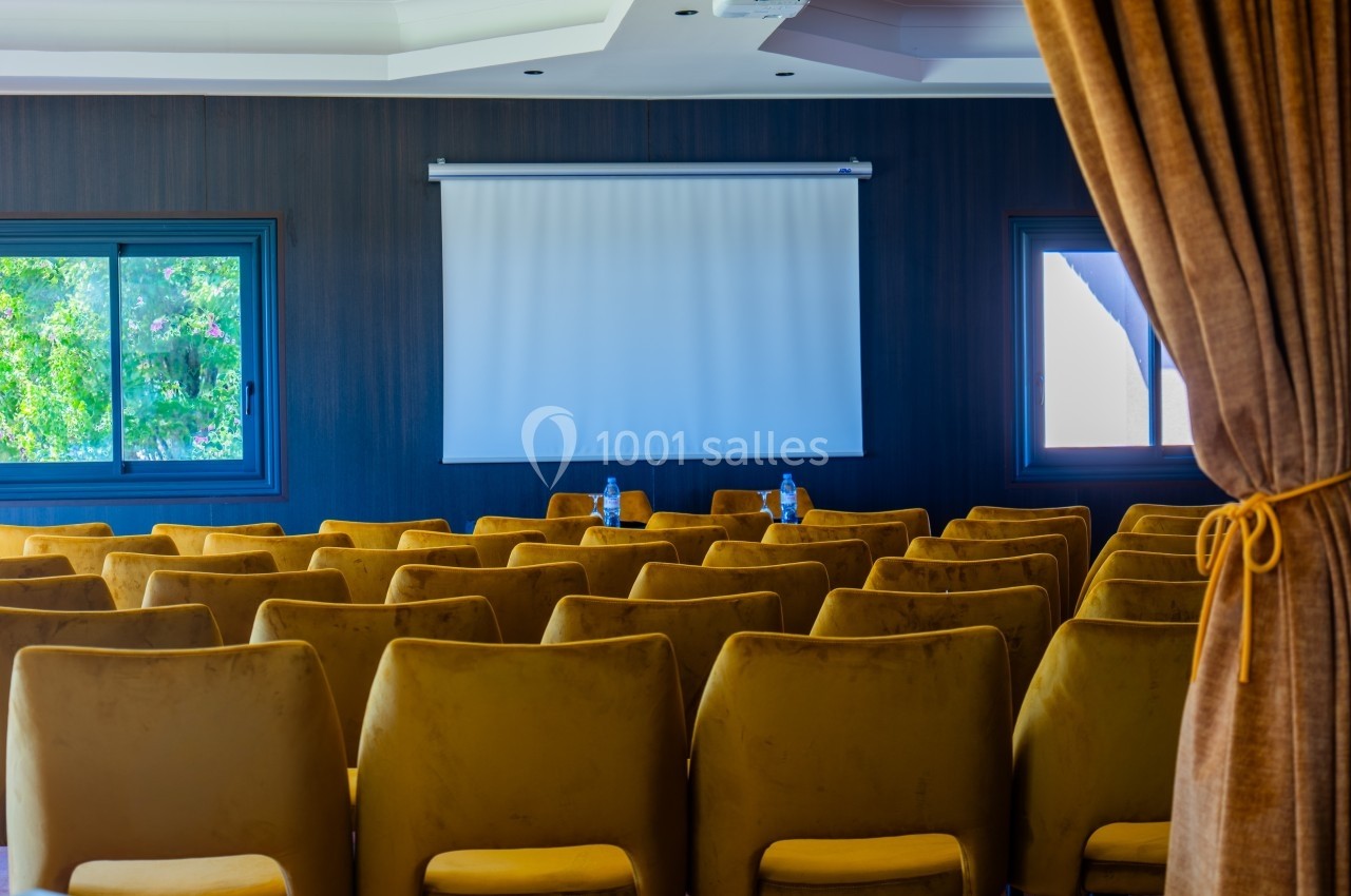 Salle de conférence avec des rangées de chaises jaunes, un écran de projection blanc et des fenêtres donnant sur l'extérieur.