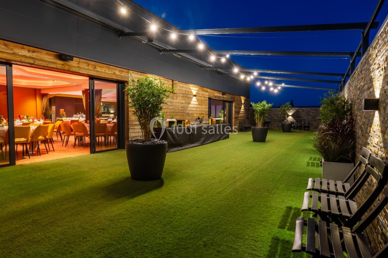 Terrasse extérieure avec pelouse artificielle, éclairage suspendu et vue sur une salle de réception illuminée.