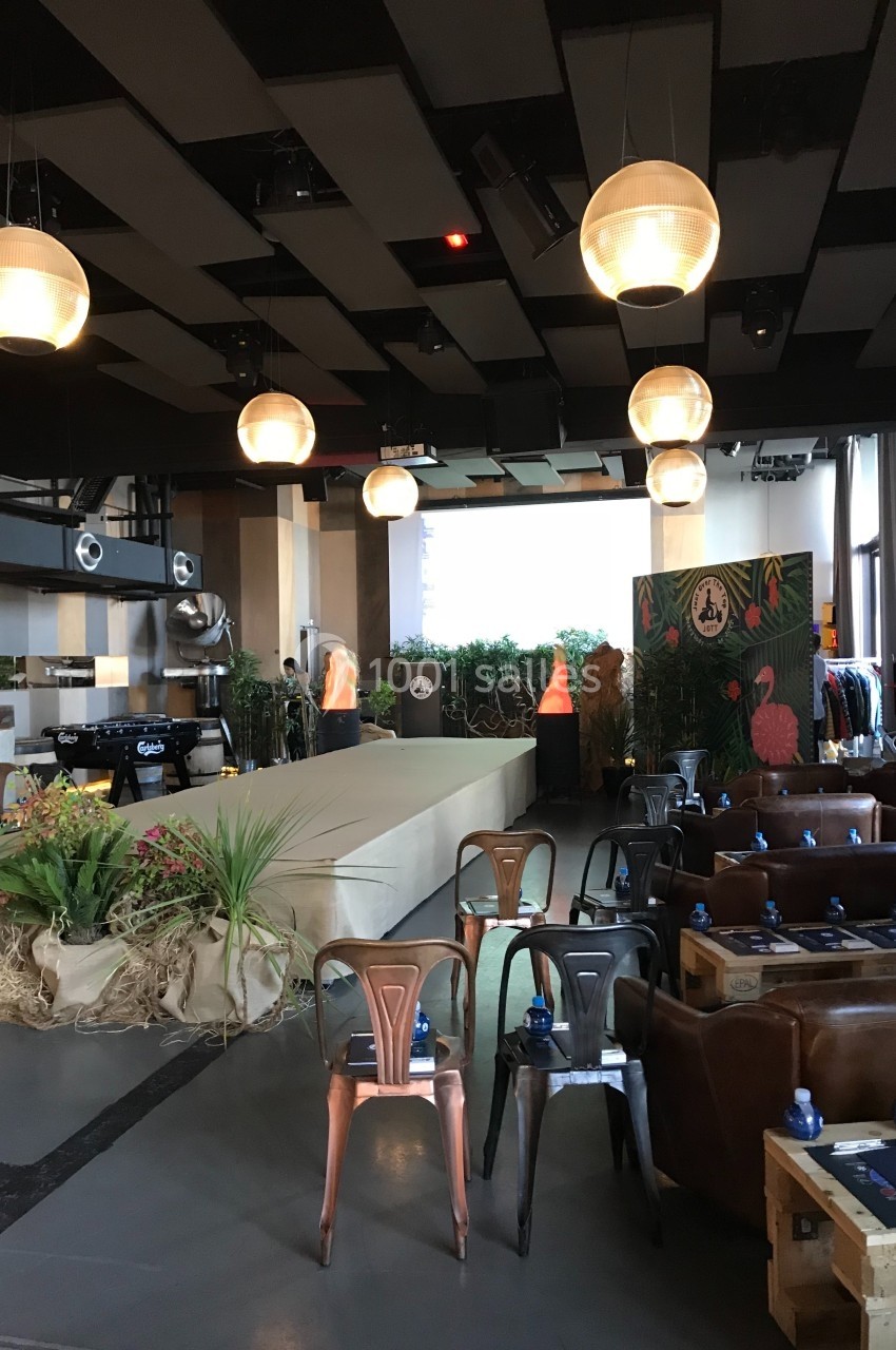 Salle aménagée avec des chaises, des tables, une scène, des plantes décoratives et des suspensions lumineuses.