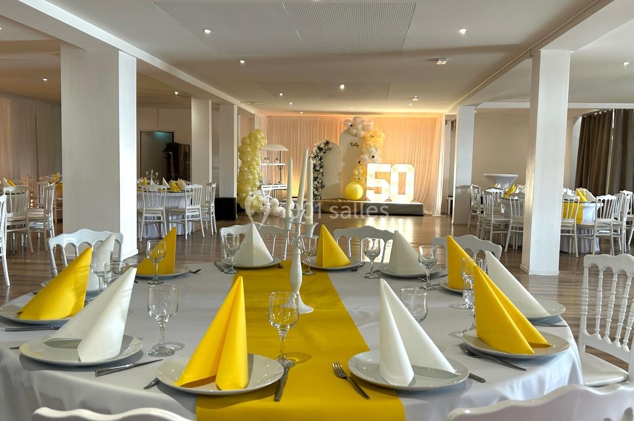 Salle décorée pour une fête avec tables dressées en jaune et blanc, et un grand ’50’ lumineux en arrière-plan.