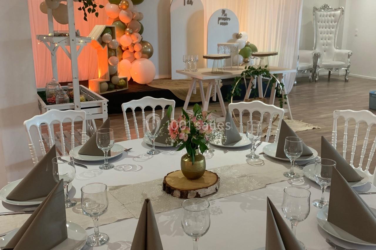 Salle décorée pour un événement avec ballons, table dressée, chaises blanches et éclairage chaleureux.
