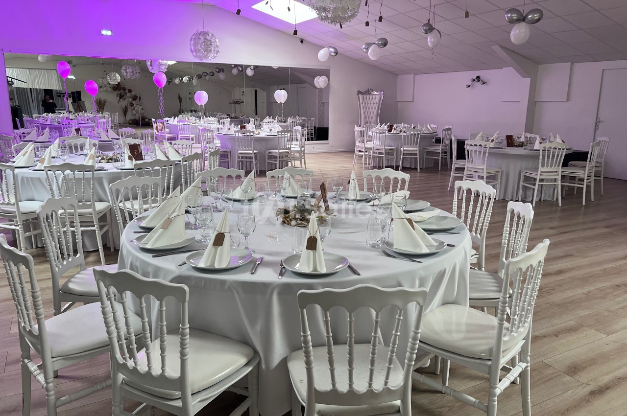 Salle de réception décorée avec des tables rondes dressées, chaises blanches et éclairage rose en arrière-plan.