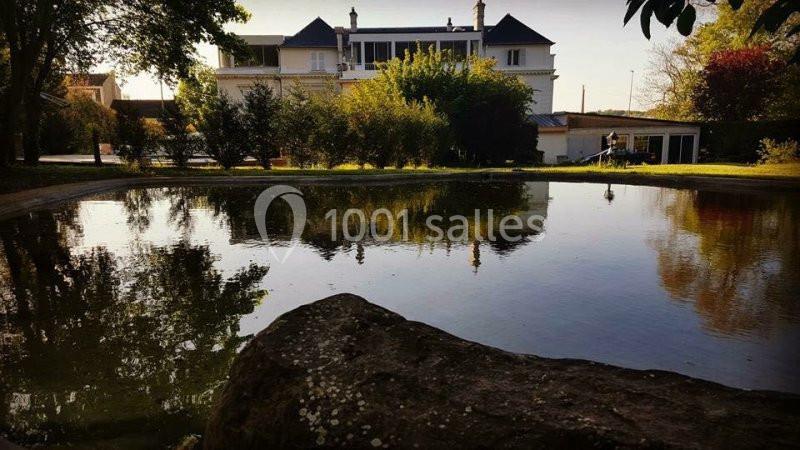Étang calme entouré de végétation, avec une grande maison blanche et des arbres reflétés dans l'eau.