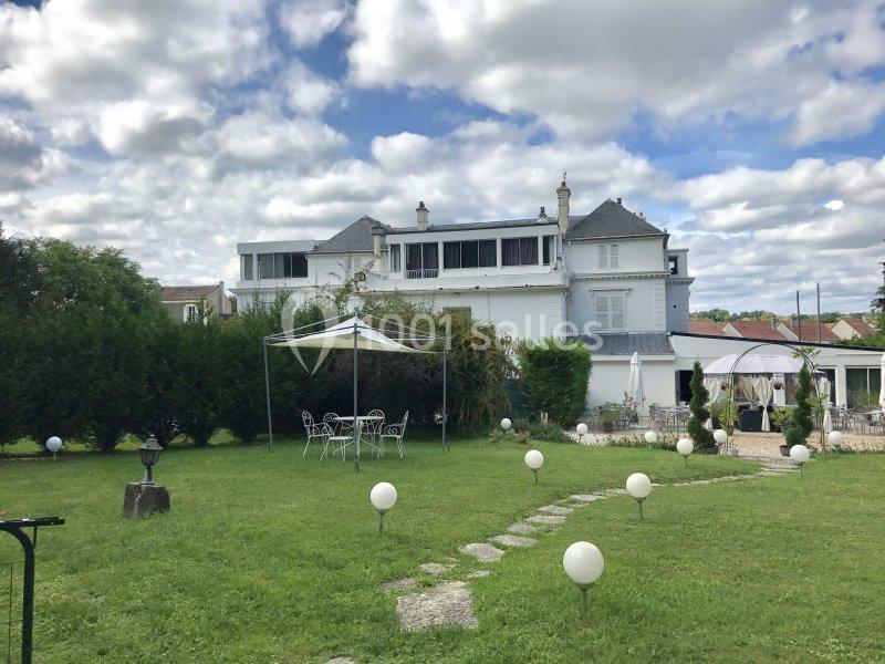 Jardin avec pelouse, chemin en pierre, mobilier de jardin sous tonnelle, et grande maison en arrière-plan.