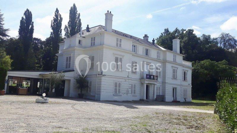 Grande maison blanche à deux étages avec volets fermés, entourée d'arbres et d'une cour en gravier.