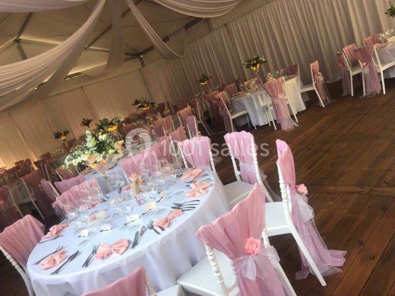 Salle de réception décorée avec des tables rondes, nappes blanches, chaises ornées de housses roses et fleurs.