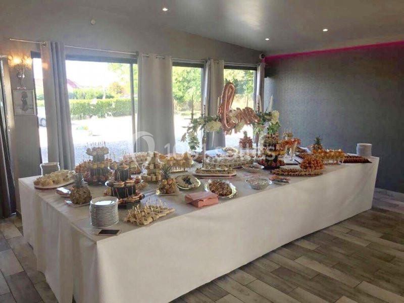 Table garnie d'un buffet varié avec amuse-bouches, desserts et décorations dans une salle lumineuse.