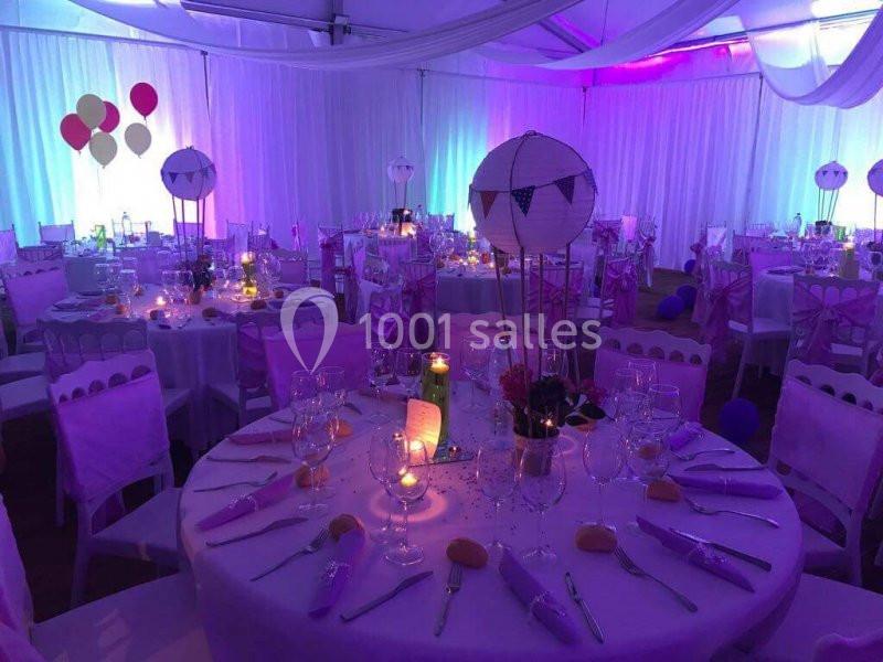 Salle de réception décorée pour un événement, avec tables dressées, éclairage coloré et ballons en arrière-plan.