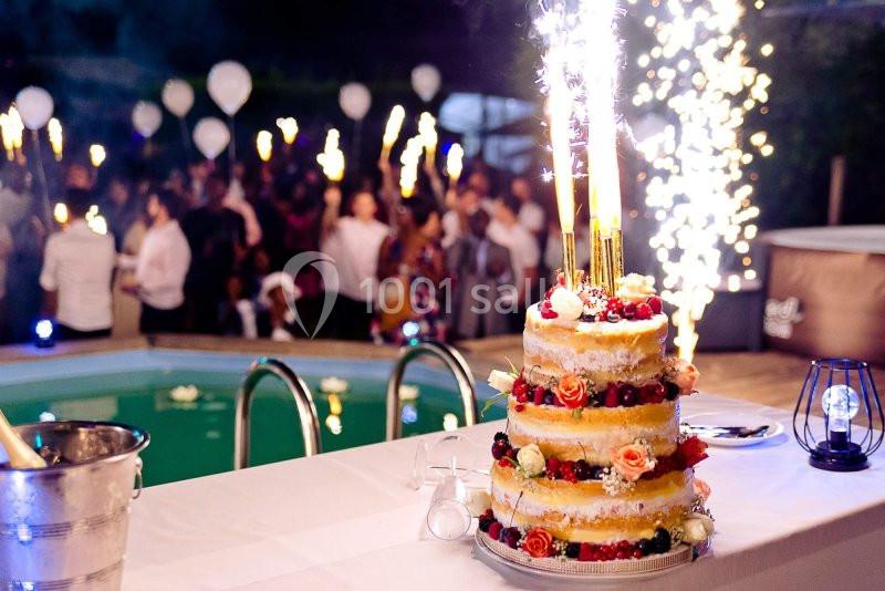 Gâteau de mariage décoré de fruits et fleurs avec des cierges magiques allumés, près d'une piscine lors d'une fête nocturne.