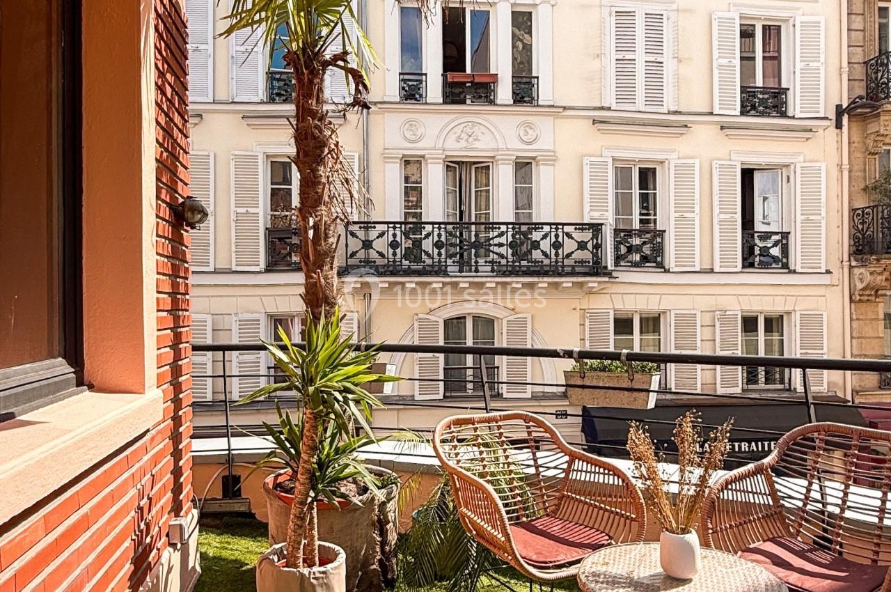 Terrasse avec gazon artificiel, mobilier en rotin, plantes et vue sur des immeubles parisiens aux volets blancs.