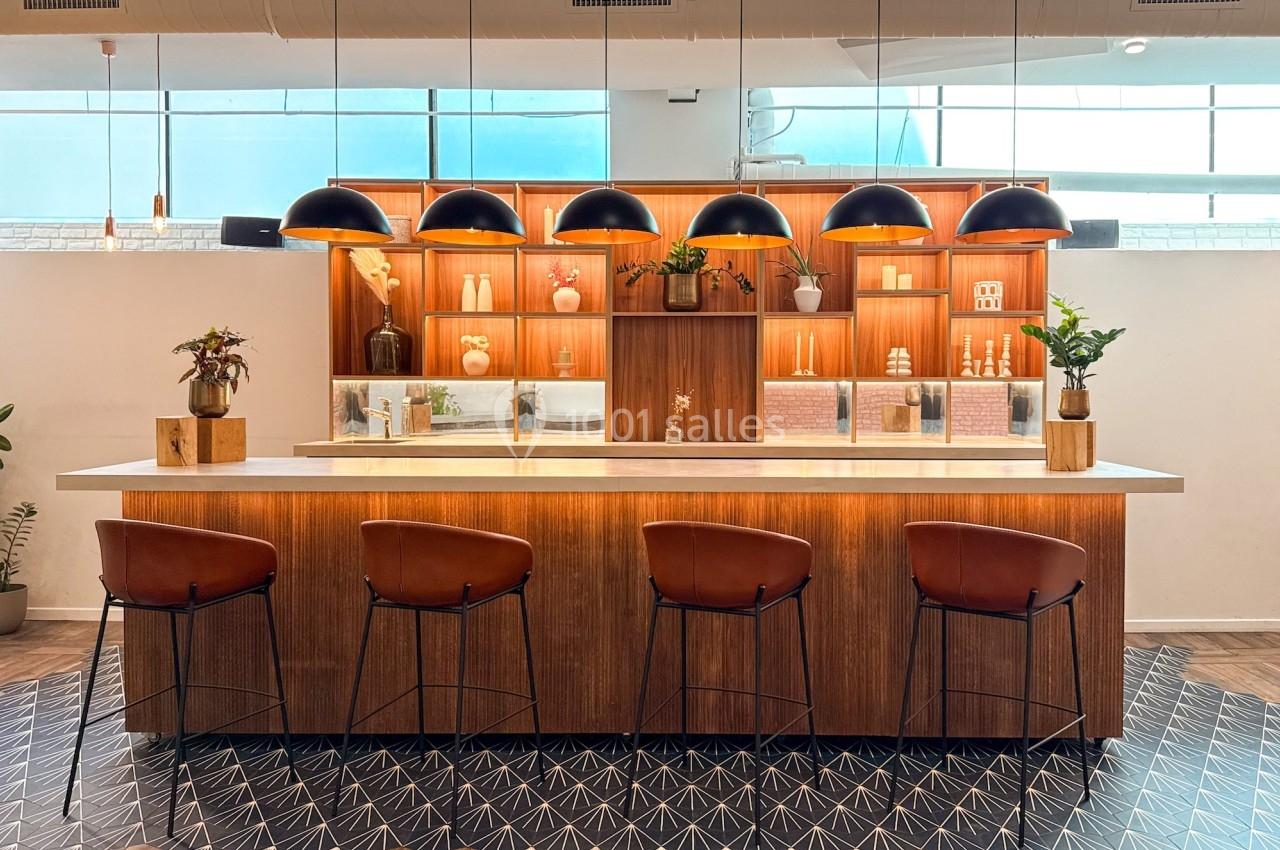 Comptoir en bois avec tabourets, étagères décoratives éclairées et suspensions noires dans un espace moderne lumineux.