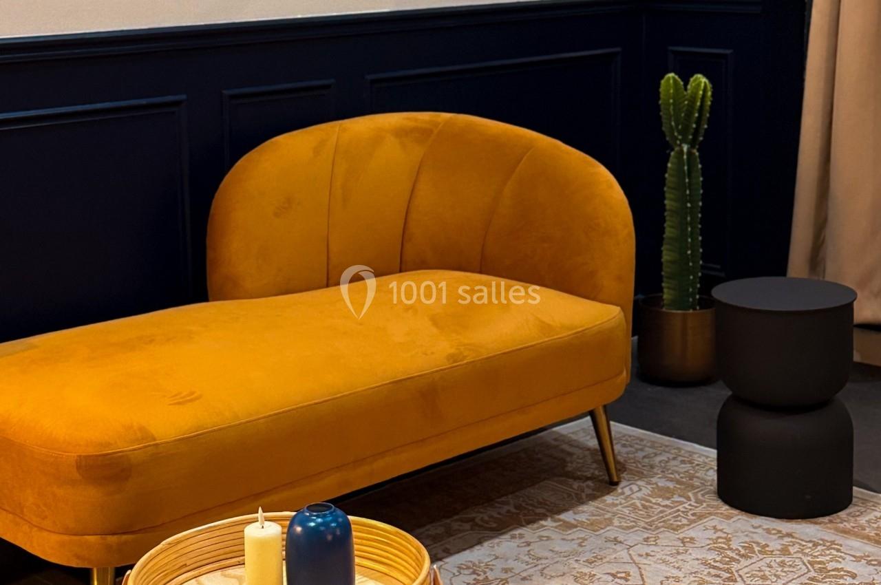 Canapé jaune moutarde sur un tapis beige, avec une table basse en rotin, un cactus et un tabouret noir en arrière-plan.