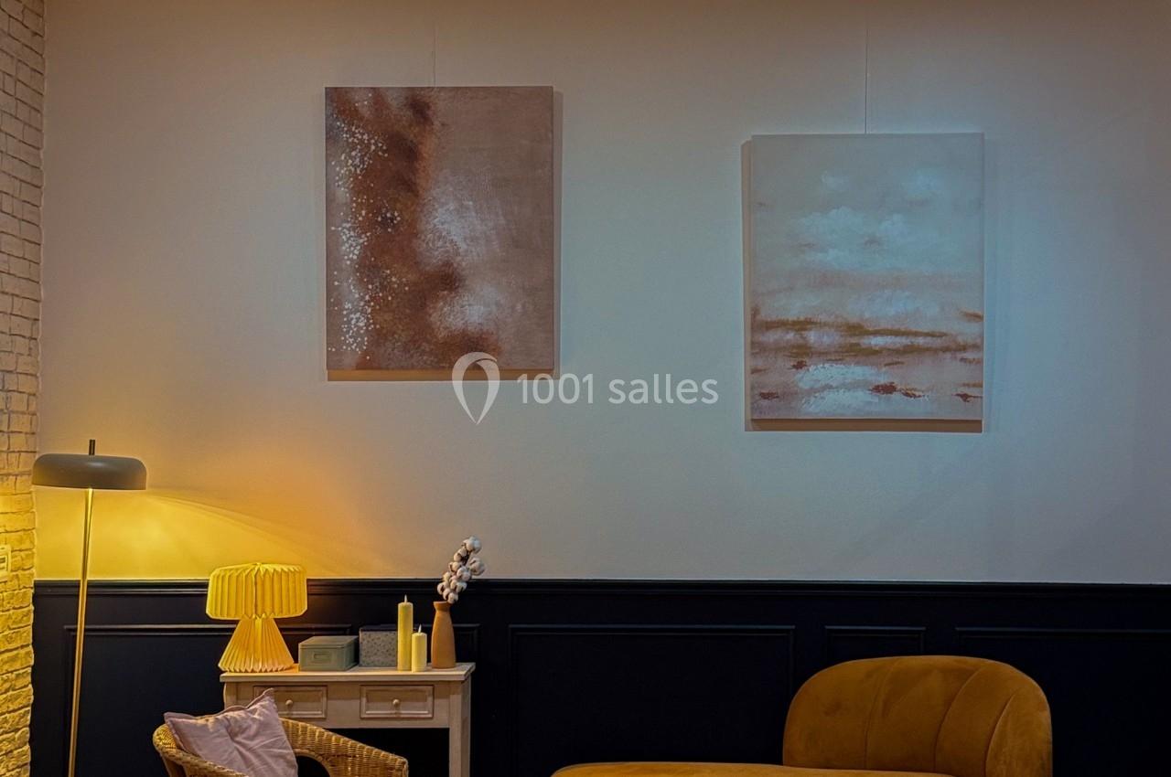 Salon moderne avec canapé jaune, fauteuils en rotin, table basse en bois, lampes et tableaux abstraits sur un mur clair.