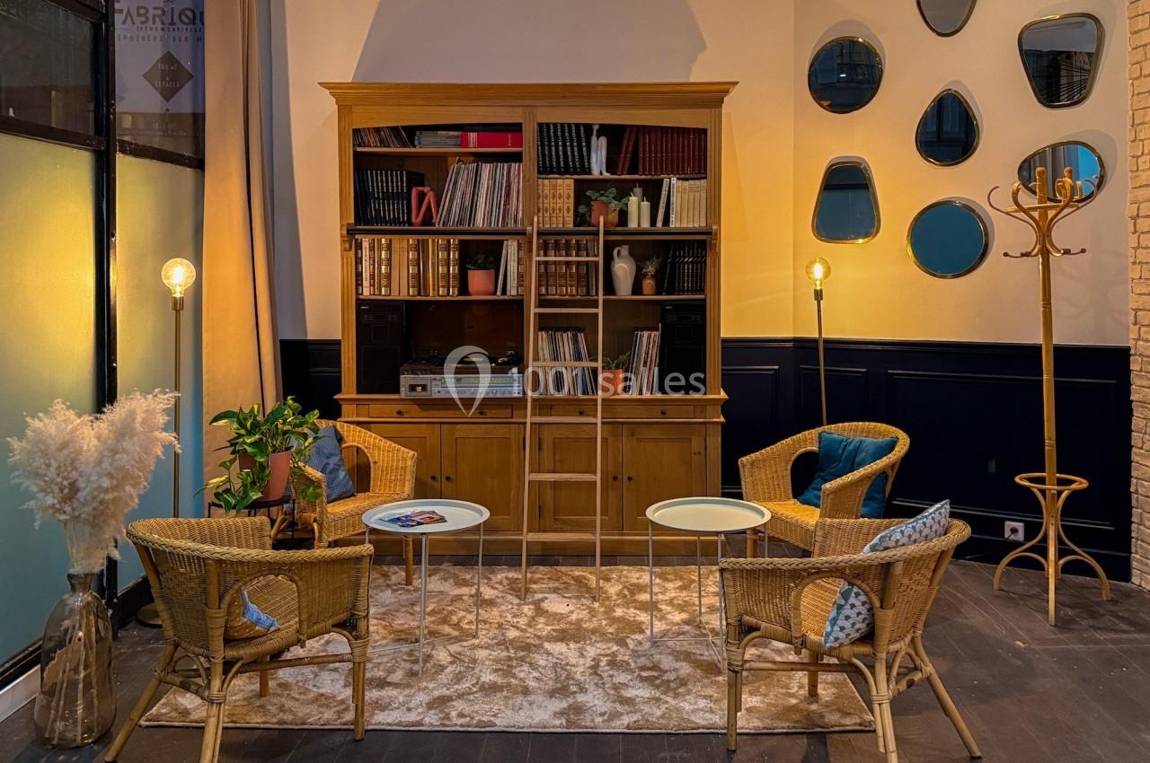 Salon chaleureux avec étagère en bois, fauteuils en rotin, tapis beige, luminaires et miroirs décoratifs au mur.