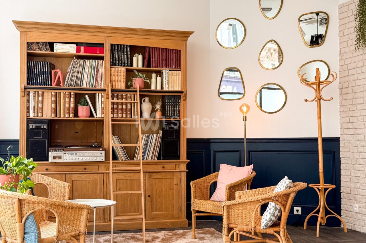 Bibliothèque en bois avec livres et vinyles, fauteuils en osier, lampadaire et miroirs décoratifs sur un mur blanc.