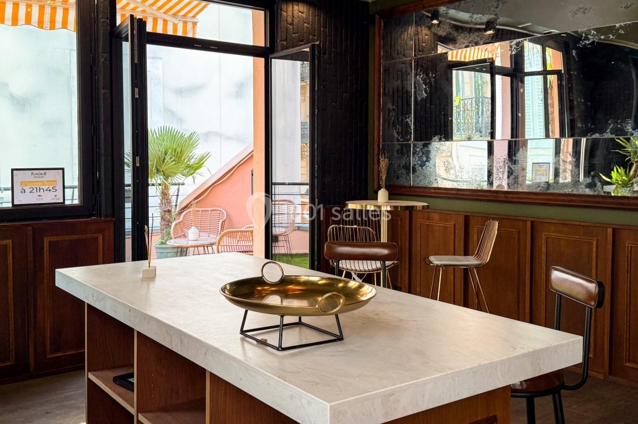Intérieur lumineux avec un comptoir en marbre, tabourets en cuir, miroir mural et vue sur une terrasse extérieure.