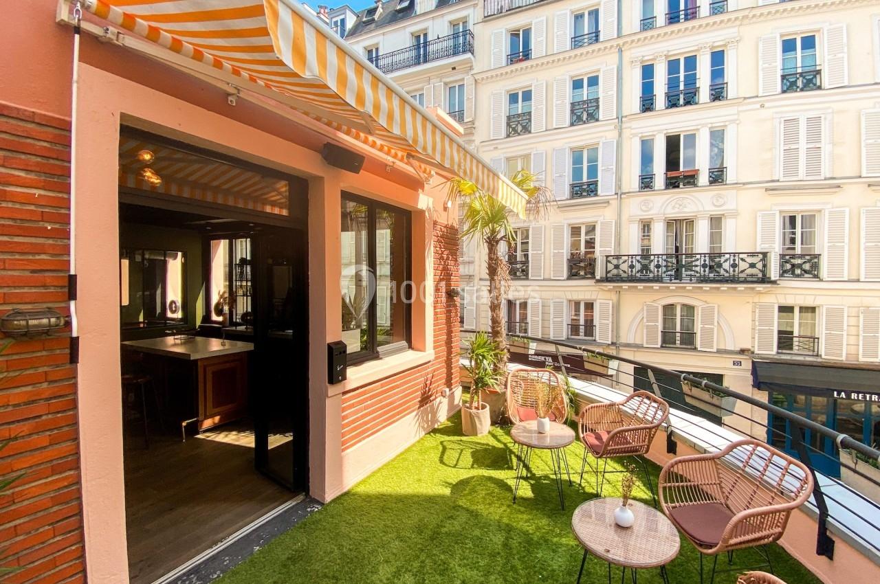 Terrasse ensoleillée avec mobilier en rotin, pelouse artificielle et vue sur des immeubles parisiens.