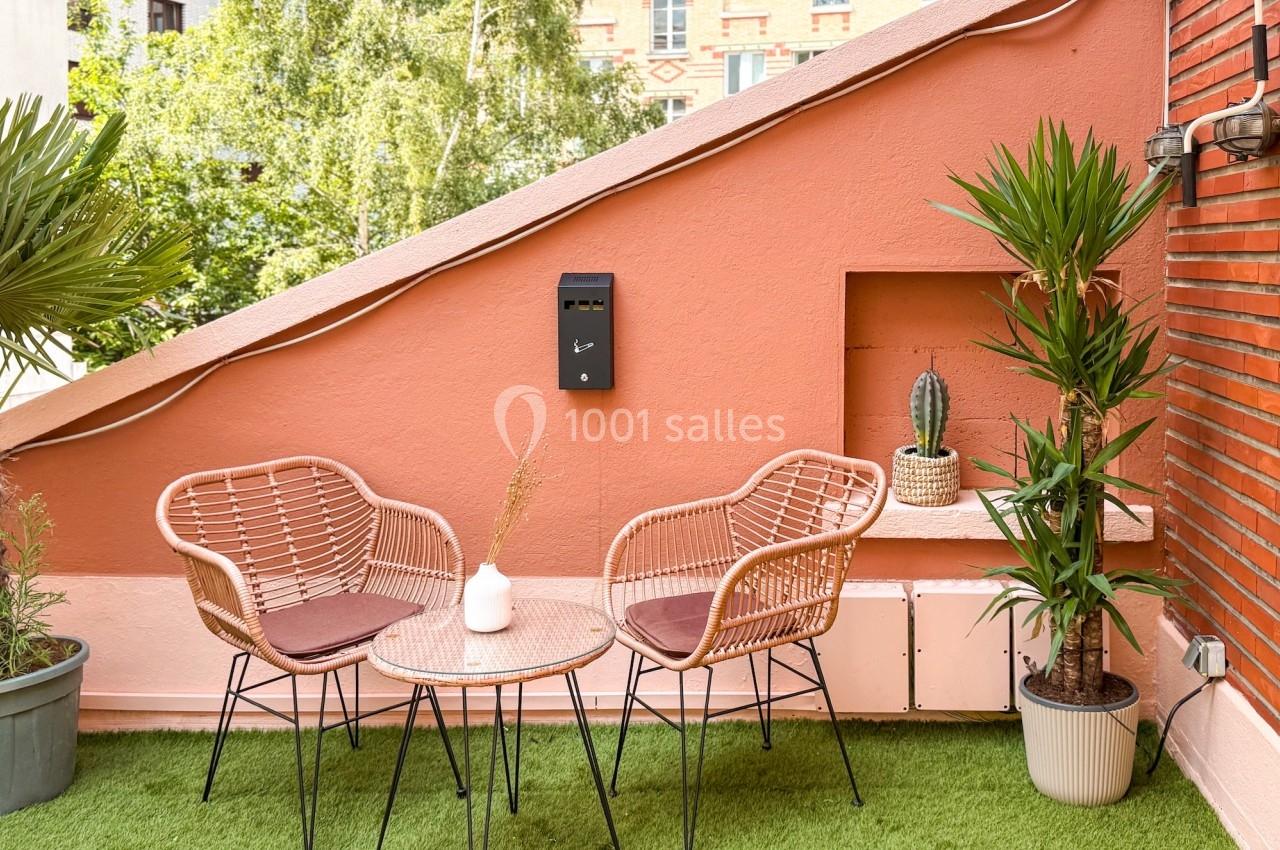 Terrasse avec table ronde, deux fauteuils en rotin, plantes en pot et mur rose, sur fond de verdure et briques.