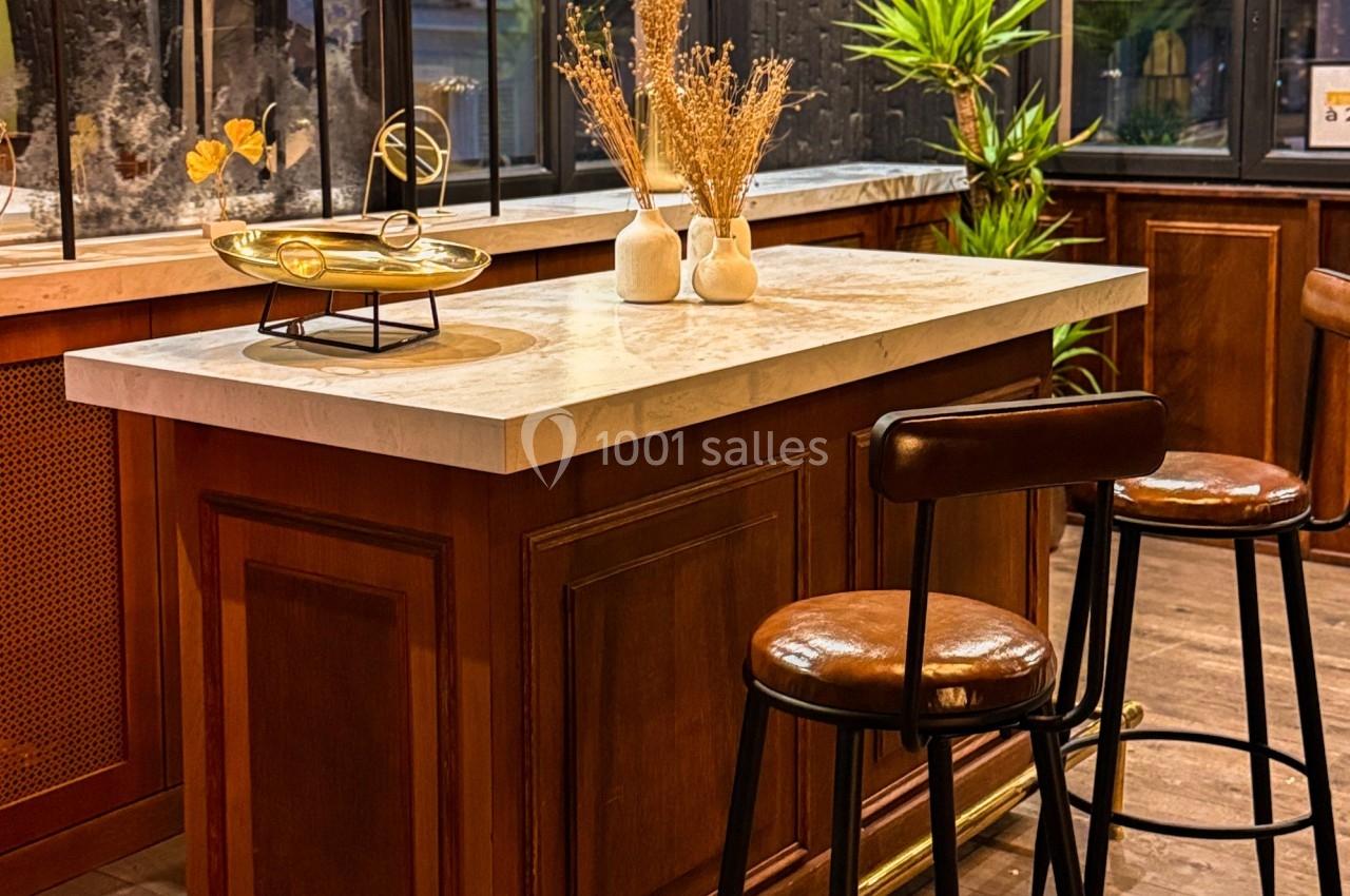 Comptoir en bois avec plateau en marbre, tabourets hauts en cuir, plantes et décoration dans un intérieur chaleureux.