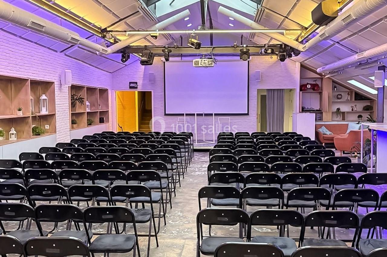 Salle de conférence lumineuse avec rangées de chaises noires, écran de projection et verrière en plafond.