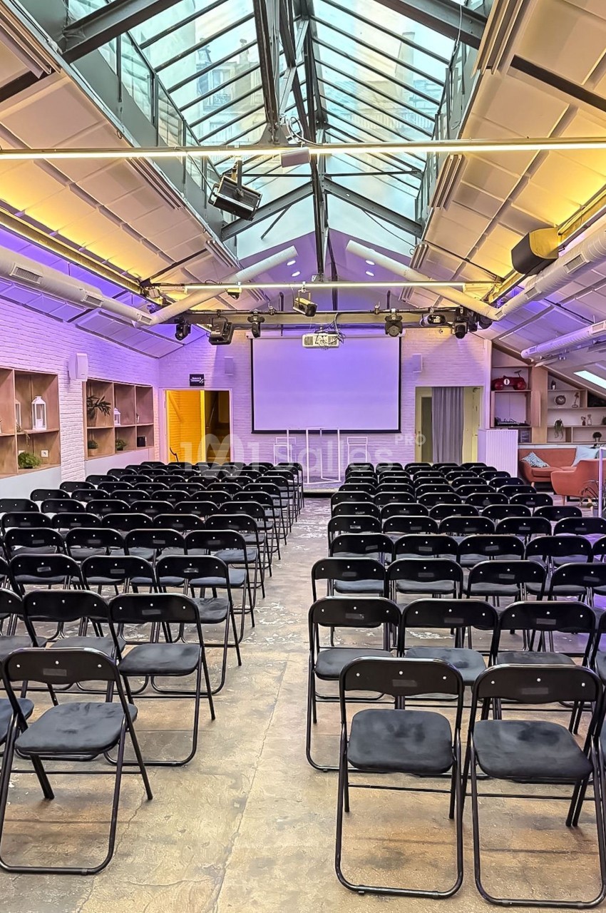 Salle de conférence lumineuse avec rangées de chaises noires, écran de projection et verrière en plafond.
